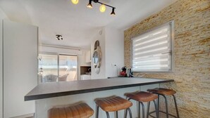 Een keuken met een centraal eiland, barkrukken, een raam met zonwering en een wand met een stenen textuur.