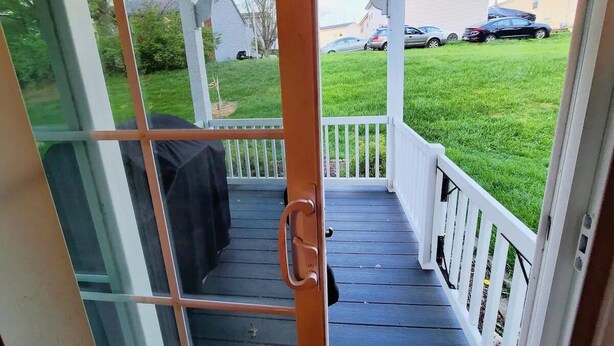 A covered porch with a wooden door, white railings, and a view of a grassy yard and parked cars.