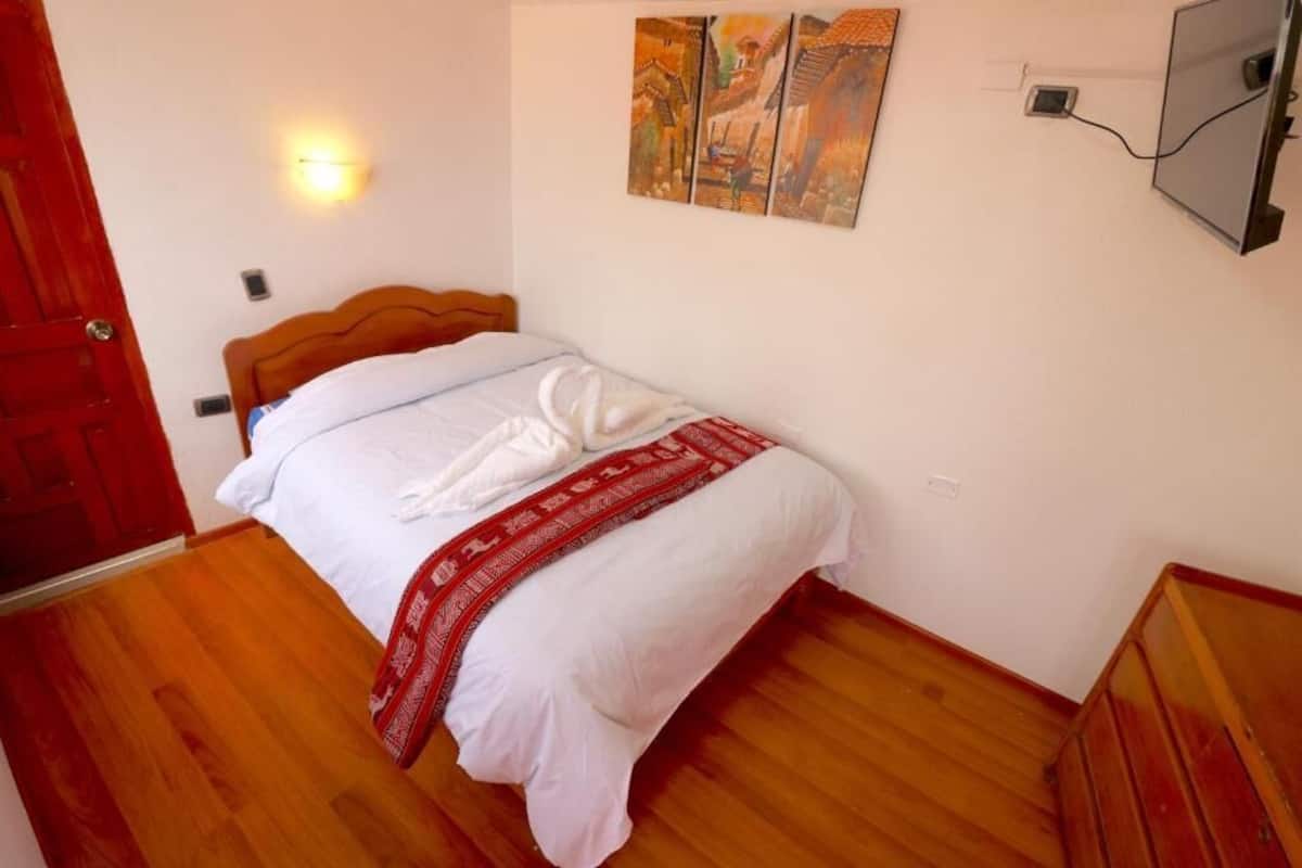 Ein Schlafzimmer mit einem hölzernen Bett, weißer Bettwäsche, einer roten Decke und einem an der Wand montierten Fernseher.