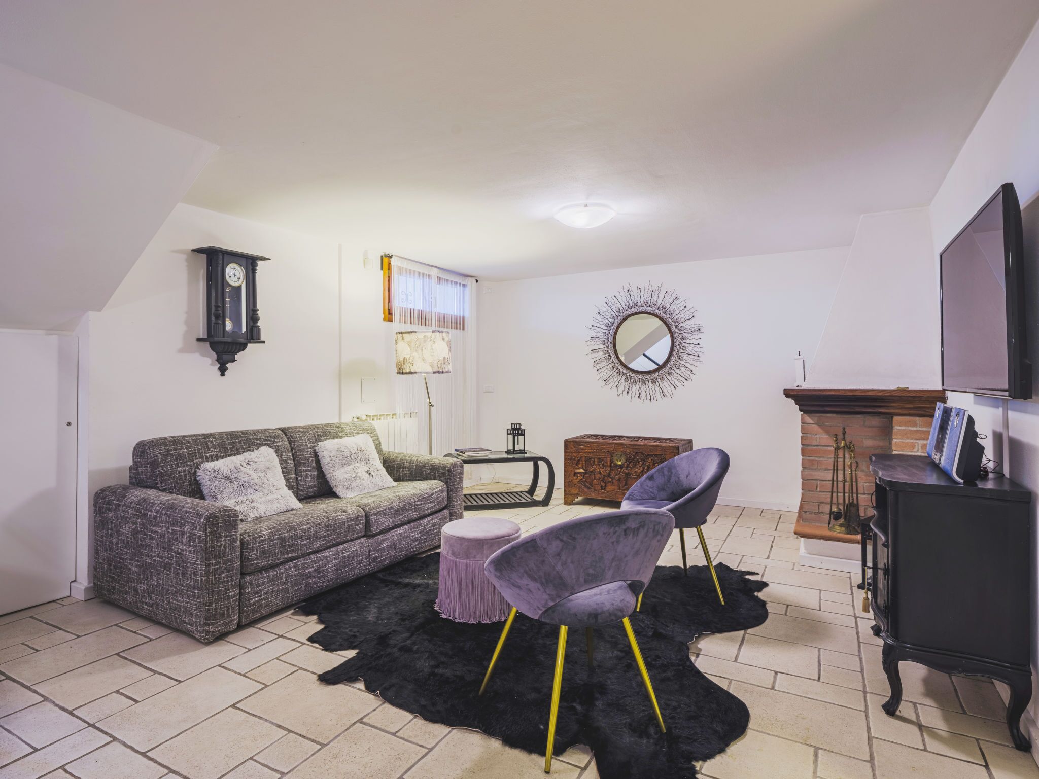 Un salon avec un canapé gris, un fauteuil violet, un tapis noir, un meuble en bois et une cheminée.