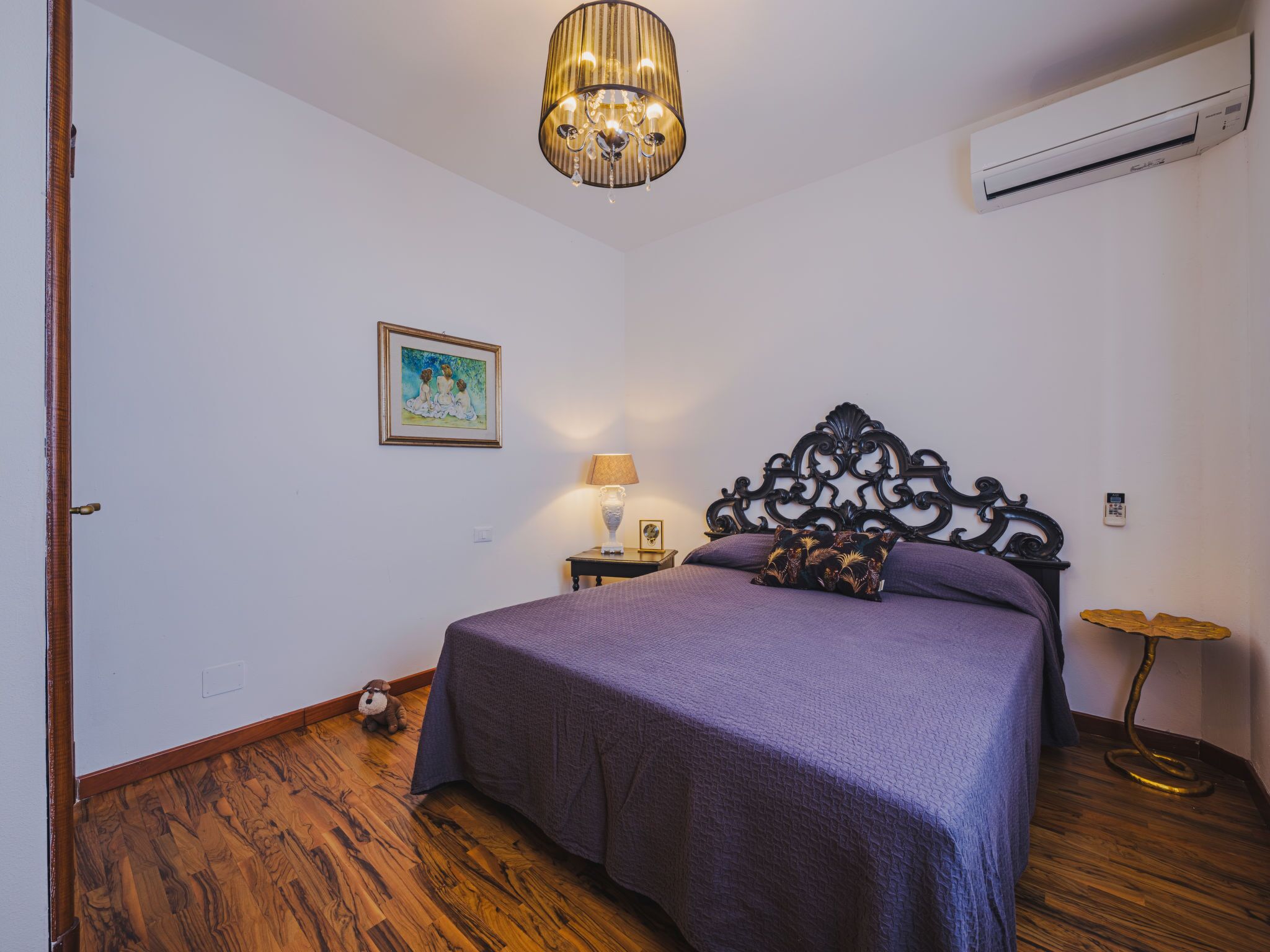 A bedroom with a bed, a nightstand, a lamp, a wall-mounted air conditioner, and a wooden floor.