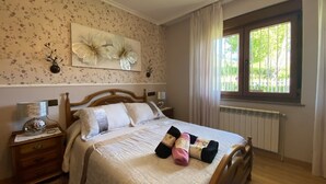 A bedroom with a wooden bed, a nightstand, a wall-mounted lamp, a radiator, and a window with curtains. - Vivienda Vacacional La Casa Azul ll (Ceceda)