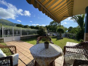 Une terrasse extérieure aménagée avec des meubles en osier, une table ronde recouverte d’une nappe à motifs floraux, et une vue sur un jardin clos avec une fontaine et des chaises.