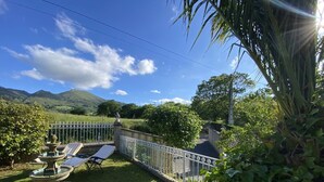 A garden with a fountain, a white fence, and a view of hills in the background. - Vivienda Vacacional La Casa Azul ll (Ceceda)