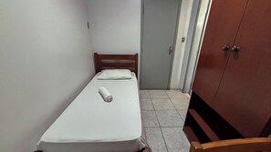 Una habitación pequeña con una cama individual, cabecera de madera, colcha blanca y una toalla enrollada sobre la cama.