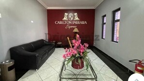 A hotel lobby with a black leather sofa, a glass coffee table with pink flowers, and a red wall with the Carlton Imbira Express logo. - Carlton Imbiara Express (Araxá)