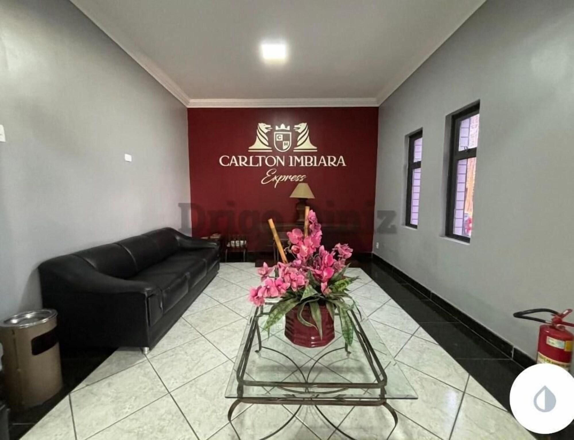 Um lobby de hotel com um sofá de couro preto, uma mesa de centro de vidro com flores cor-de-rosa e uma parede vermelha com o logo Carlton Imbira Express.