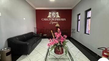 Um lobby de hotel com um sofá de couro preto, uma mesa de centro de vidro com flores cor-de-rosa e uma parede vermelha com o logo Carlton Imbira Express.