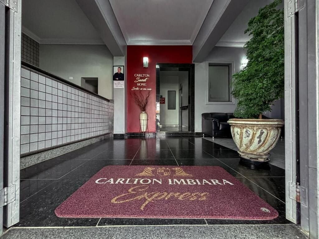Entrada de hotel con una pared roja, una planta grande en maceta y un felpudo que dice "Carlton Imbira Express".