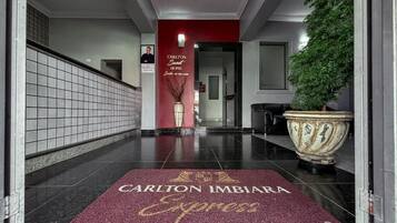 Entrada de hotel con una pared roja, una planta grande en maceta y un felpudo que dice "Carlton Imbira Express".
