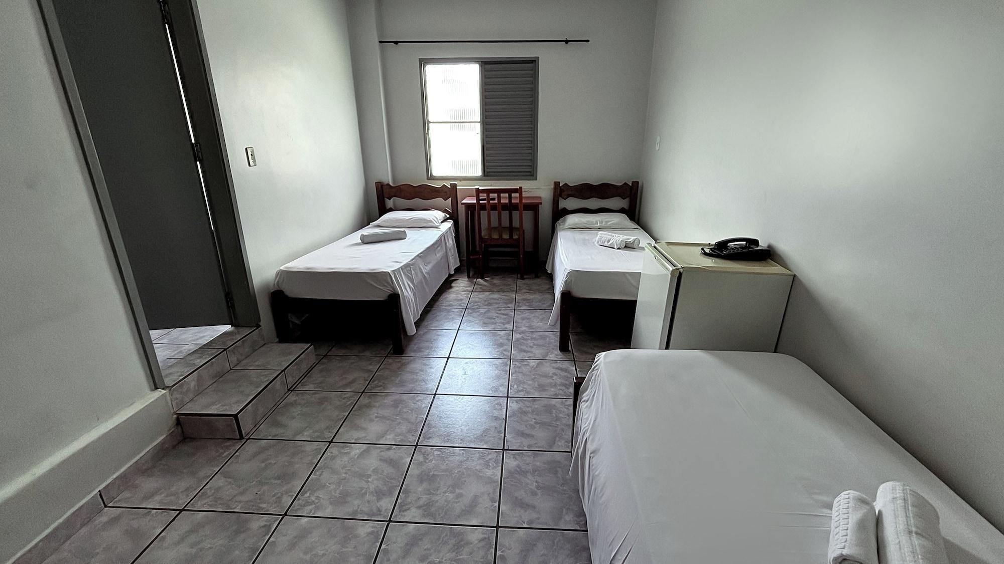 Una habitación pequeña y estrecha con dos camas individuales, un escritorio con silla y una ventana con persianas.