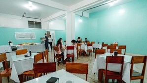 A dining area with tables and chairs, a kitchen area, and a person serving food. - Hotel Verde Mares (Macapa)