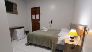 A small, clean room with a bed, a bedside table with a lamp, a wooden headboard, a small desk with a computer, and a door with a sign. - Hotel Verde Mares (Macapa)