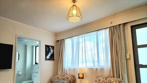 Een woonkamer met twee fauteuils met bloemenprint, een bijzettafeltje met een lamp, een groot raam met dunne gordijnen en uitzicht op de badkamer.