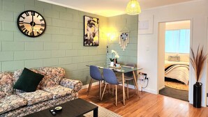 Een woonkamer met een bank, salontafel en een eethoek met stoelen en een tafel. Een wandklok en een ingelijst kunstwerk zijn zichtbaar.