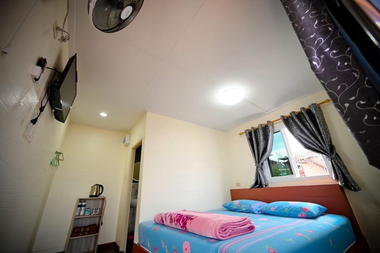 A bedroom with a bed, a TV mounted on the wall, a ceiling fan, and a window with curtains.