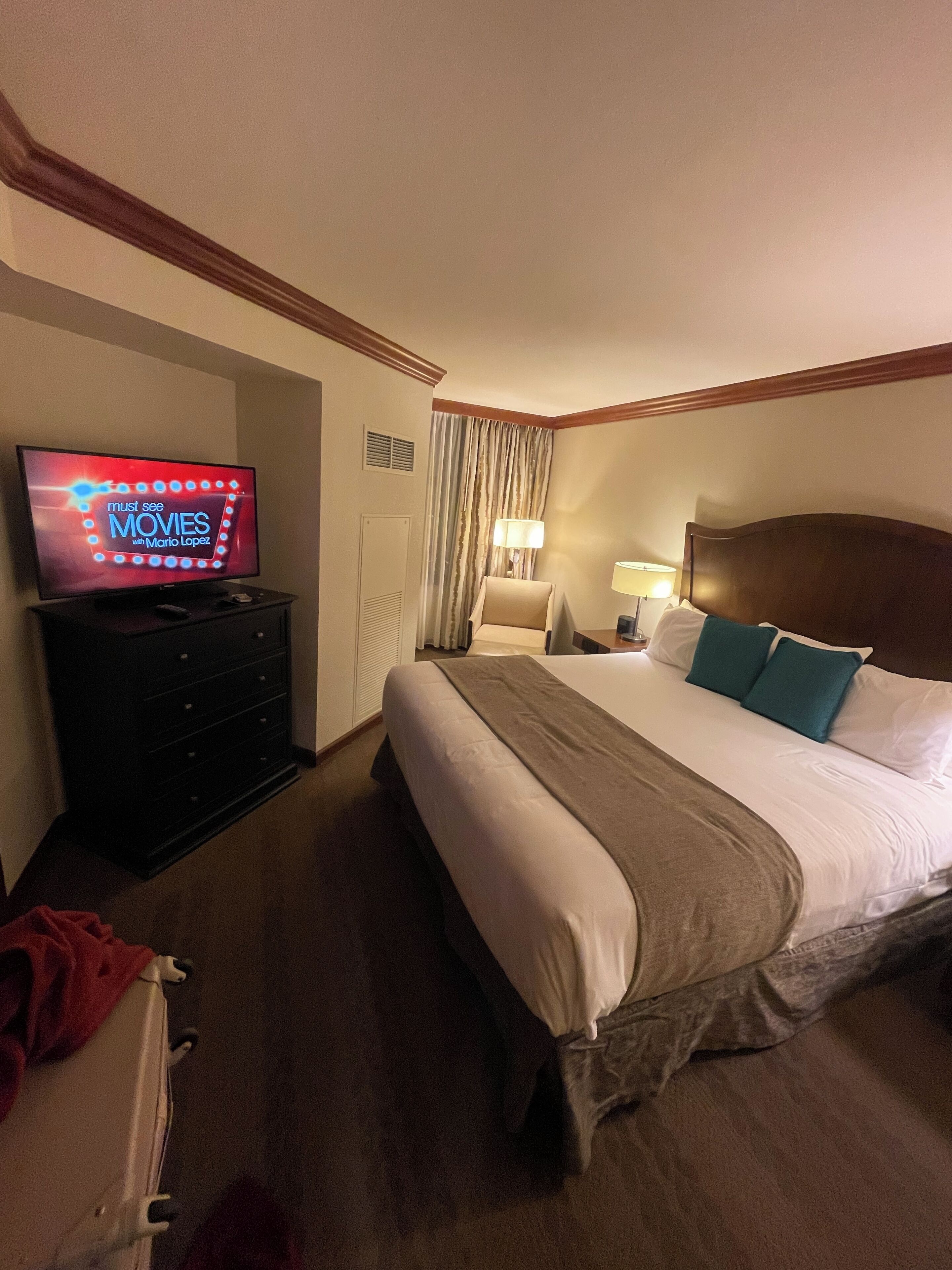 A hotel room with a bed, a TV on a dresser, a lamp, and a window with curtains.