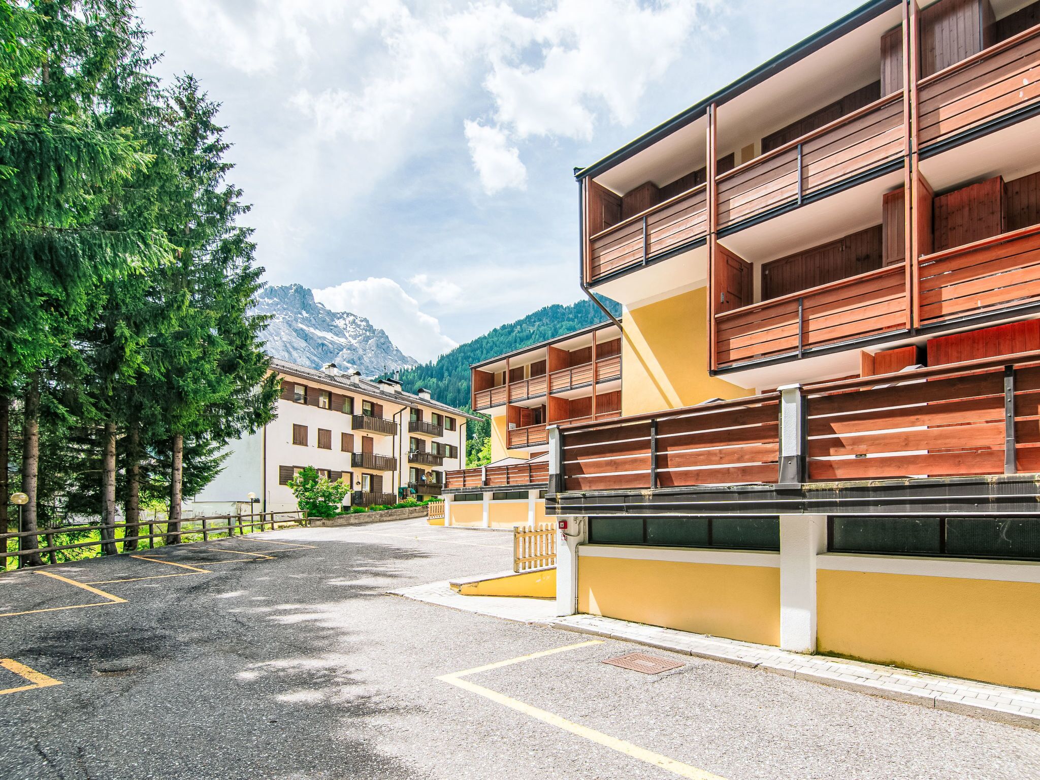 A multi-story building with balconies, a parking area, and a mountain backdrop.
