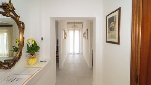 Un corridoio con uno specchio, un vaso con fiori, quadri alle pareti e una porta a destra.