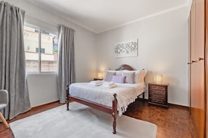A bedroom with a wooden bed, a nightstand with a lamp, a chair, and a view of the outside through a window with curtains. - Glimpse of the City by Homie (Funchal)
