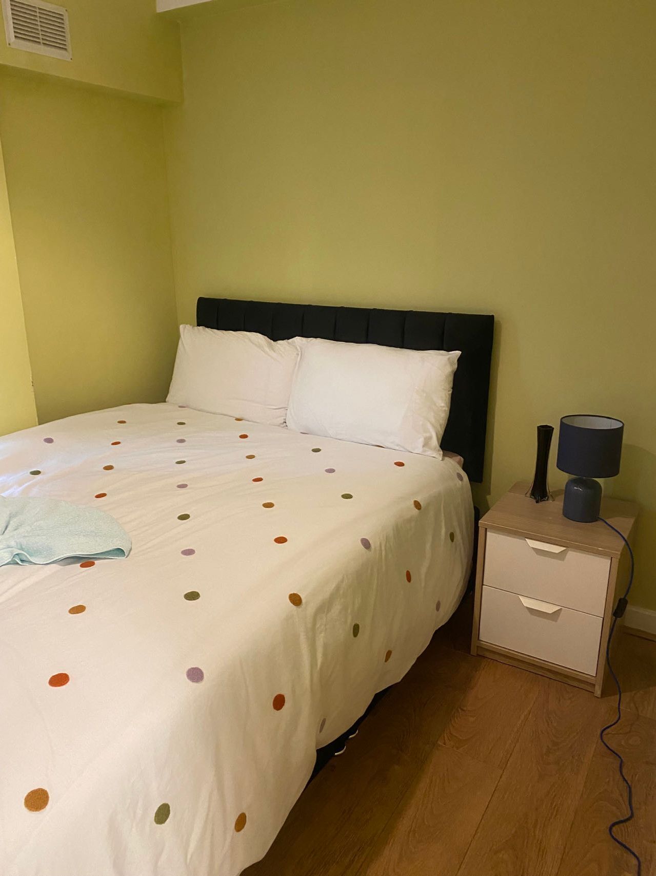 A bed with a polka dot bedspread, a bedside table with a lamp, and a wall-mounted air vent.