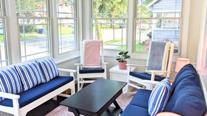 A sunroom with a view of a residential street, featuring a blue-striped sofa, a white armchair, and a small table.