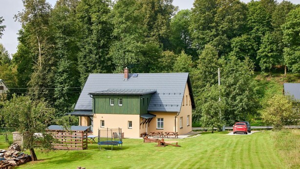 Une maison avec un toit en pente, une cour clôturée et une voiture rouge garée dans l’allée.