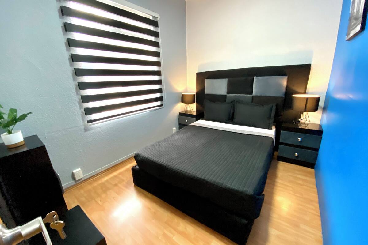 A bedroom with a bed, bedside tables, a lamp, a potted plant, and a window with blinds.