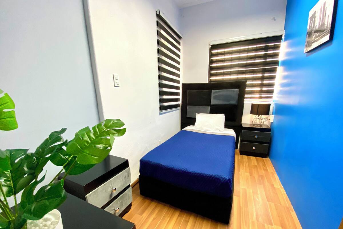 A bedroom with a bed, a nightstand, a dresser, a potted plant, and a window with blinds.