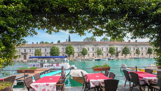 Un espace de restauration en plein air, avec des tables et des chaises, donnant sur un canal où naviguent des bateaux, et avec un bâtiment historique en arrière-plan.