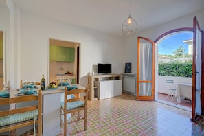 A room with a dining table set for two, a TV, a wooden chair, a small cabinet, and a view through an open door.