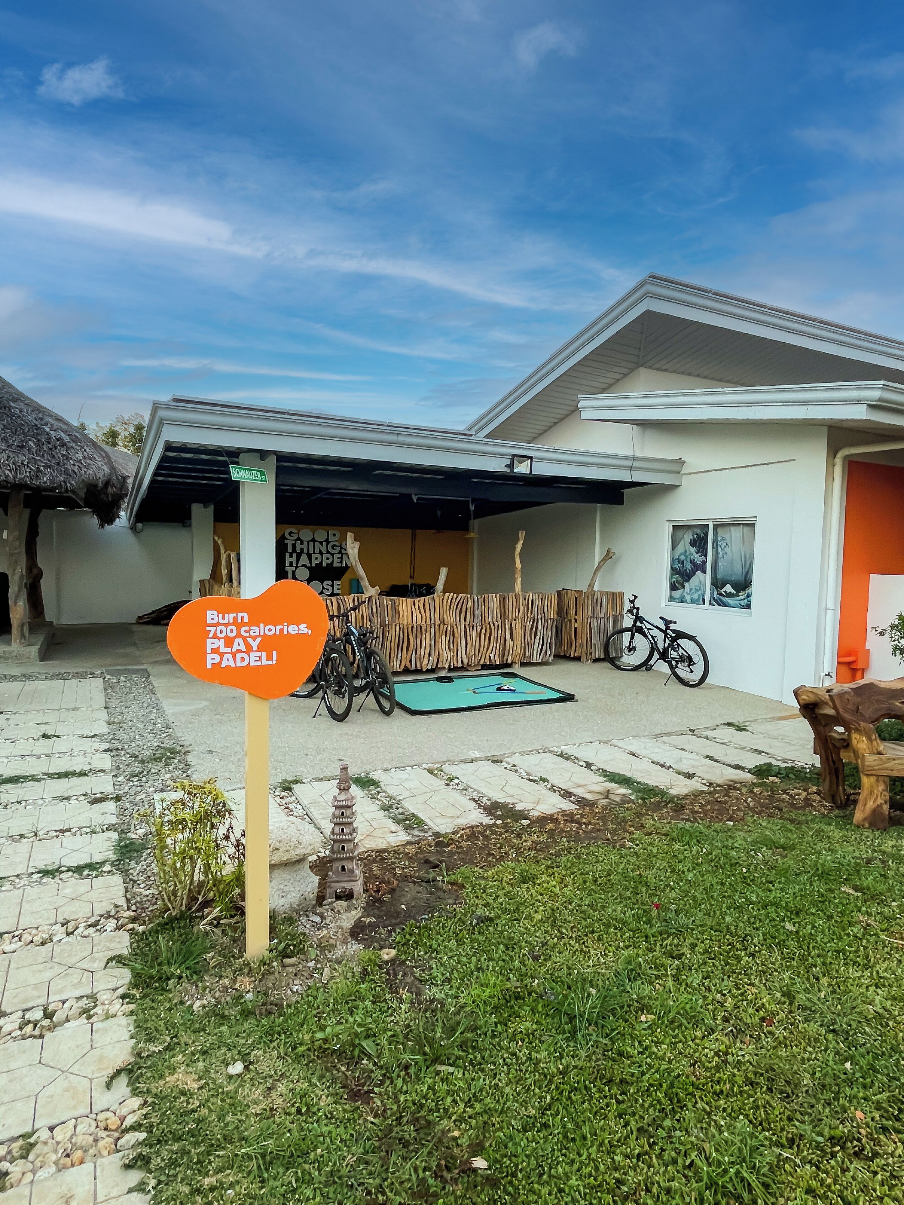 A house with a covered porch, bicycles, and a sign that reads "Burn 700 calories, PLAY PADEL!"