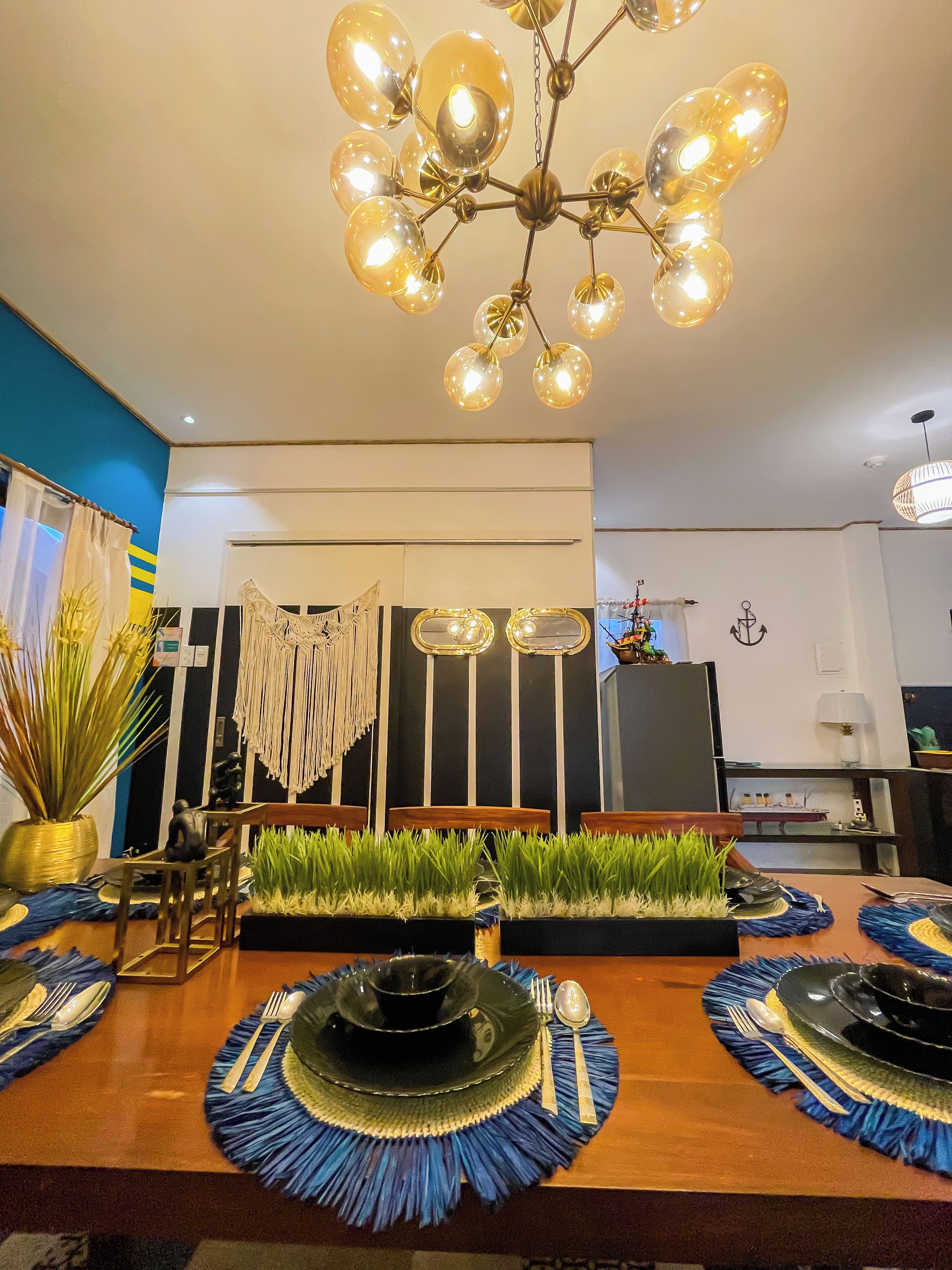 Un coin repas avec une table en bois, dressée pour quatre personnes, entourée de chaises. Un mur décoratif orné de rayures verticales et une œuvre en macramé complètent l’ensemble.