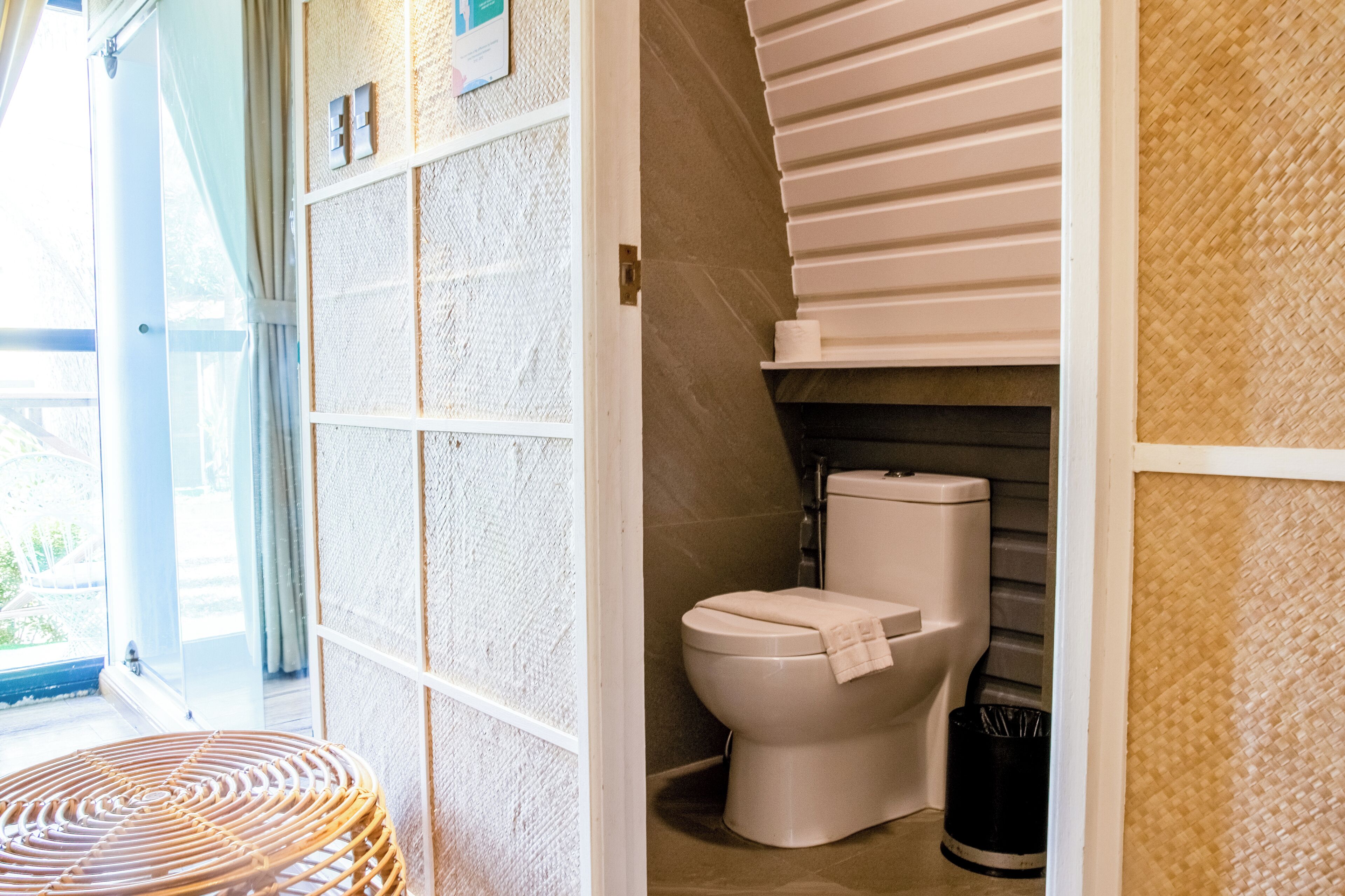 A bathroom with a toilet, a wicker chair, and a view of the outdoors.
