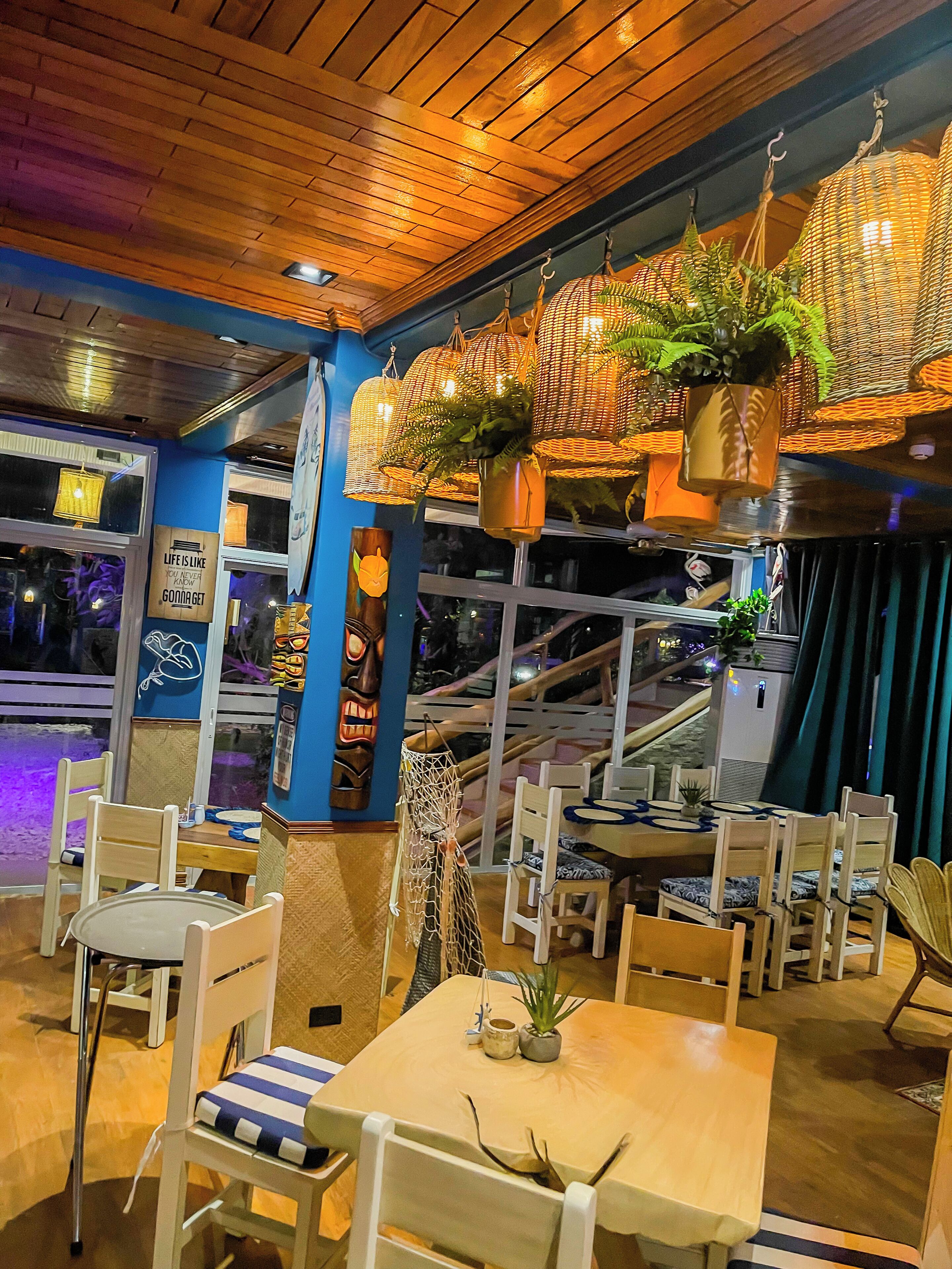 A restaurant interior with wooden ceiling, hanging wicker lamps with plants, and a view of a staircase outside.