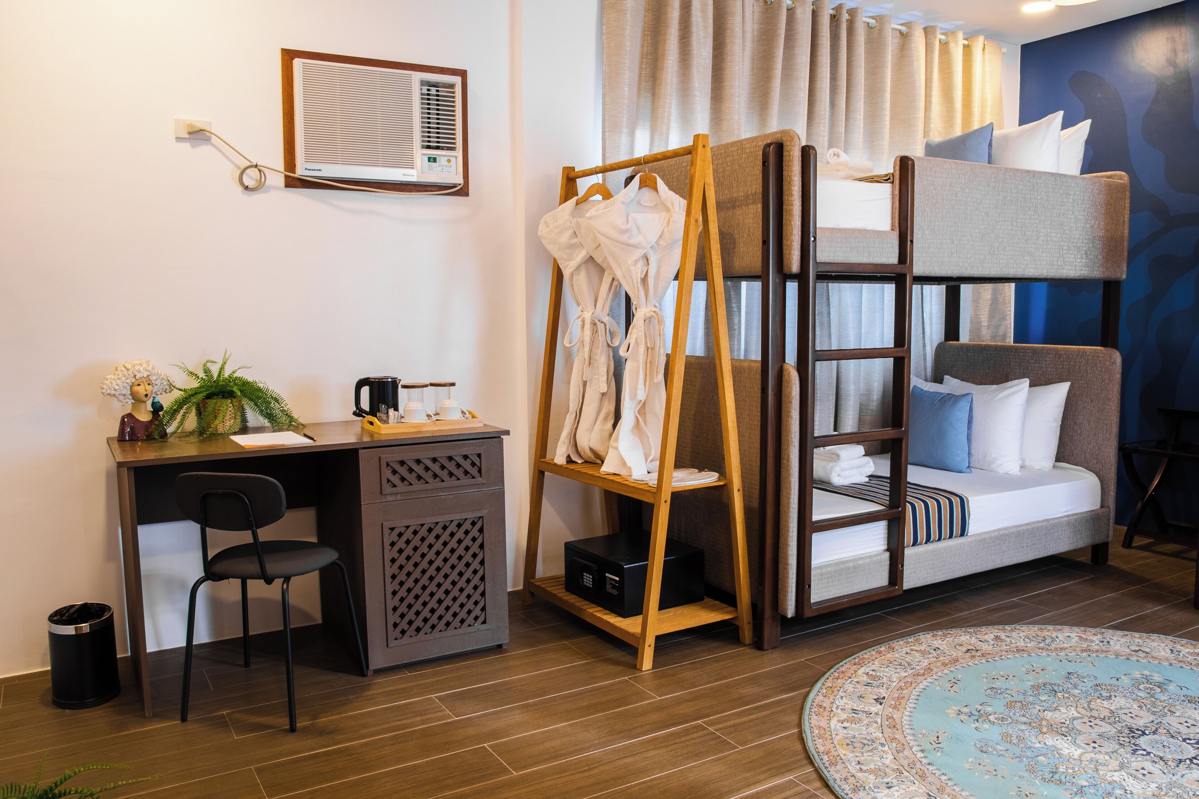 A compact hotel room with a bunk bed, a desk with a chair, a small plant, and a round rug.