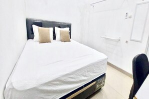 Cama bem arrumada com colchão branco e duas almofadas, cabeceira escura e porta branca com puxador.