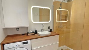 Casa de banho moderna com toucador de madeira, lavatório branco, espelho com luz, chuveiro manual e máquina de lavar roupa.