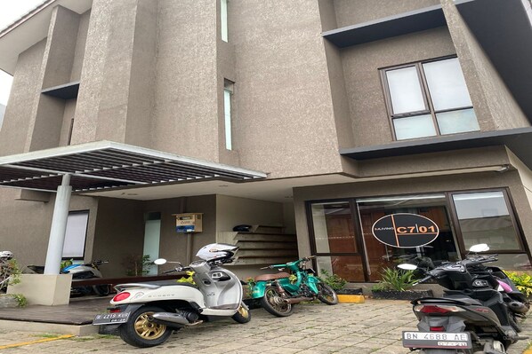 A modern building with a covered entrance, a sign with a circular logo, and several parked scooters and motorcycles. - OYO 93998 Three J Living (Curug)