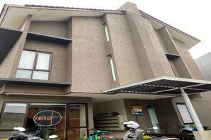A modern building with a large glass entrance, a visible logo, and a covered outdoor area with parked scooters. - OYO 93998 Three J Living (Curug)