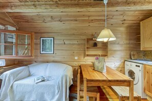 Une cabane confortable avec un intérieur en bois, un lit, une table, un lave-linge et une fenêtre donnant sur les arbres.
