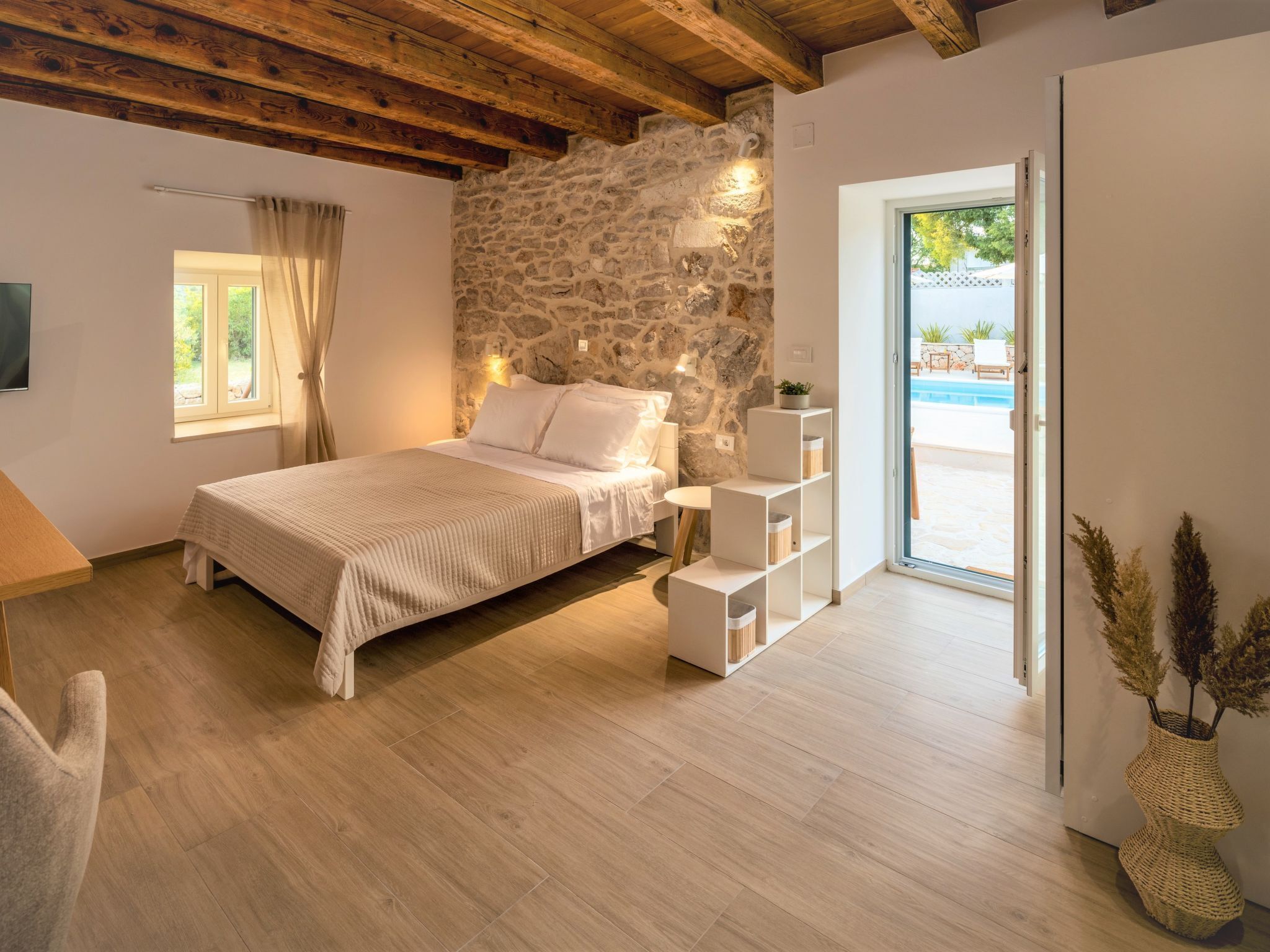 A bedroom with a stone wall, a bed, a bedside table, a lamp, a window, and a view of an outdoor swimming pool.