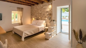A bedroom with a stone wall, a bed, a bedside table, a lamp, a window, and a view of an outdoor swimming pool. - Villa di Perla by Interhome (Primošten)