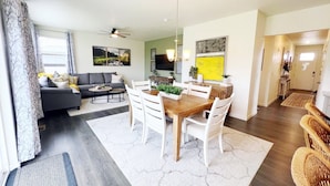 Ein modernes Wohnzimmer mit einem grauen Sofa, einem Esstisch mit weißen Stühlen, einem Deckenventilator und einem großen Fenster mit Vorhängen.