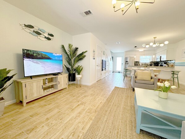 A modern living room with a flat-screen TV, wooden flooring, a large potted plant, and a dining area with a table and chairs.