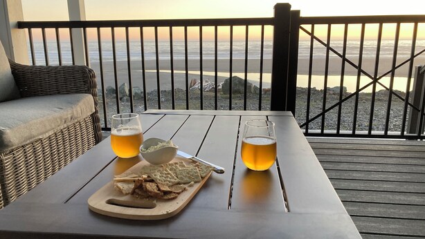 Ein Tisch mit einem Bierglas, einer Schüssel Chips und einem Schneidebrett mit Brot, mit Blick auf den Ozean auf einer Holzterrasse.