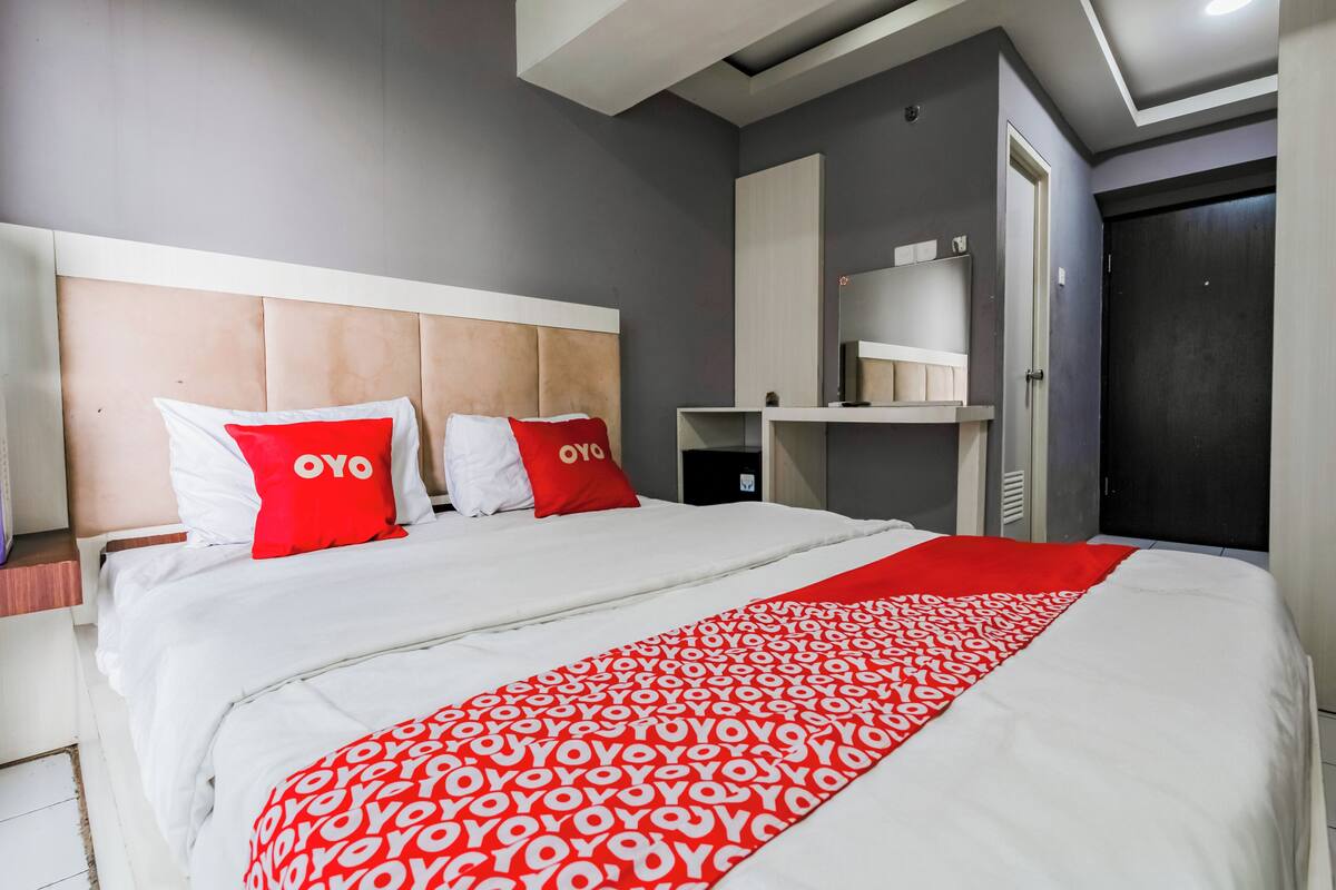 Ein modernes Hotelzimmer mit einem großen Bett, zwei roten Kissen, einer mehrfarbigen Decke in Weiß und Rot, einem Holzkopfteil, einem Nachttisch und einem Kleiderschrank.