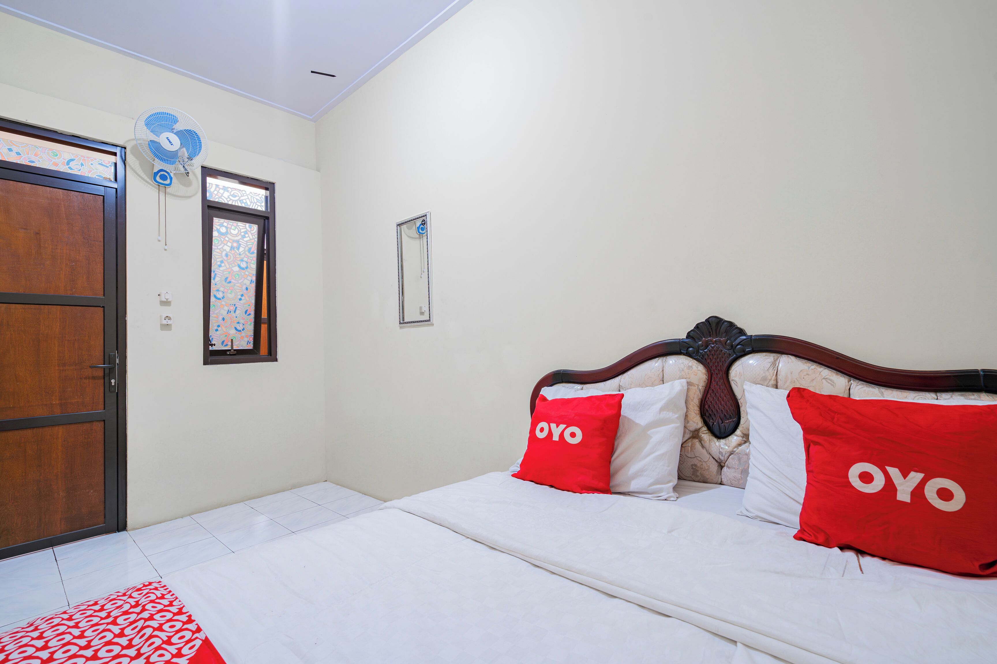 Een slaapkamer met een bed, twee rode kussens met de tekst 'OYO', een houten deur, een raam met decoratief patroon en een ventilator bevestigd aan de muur.