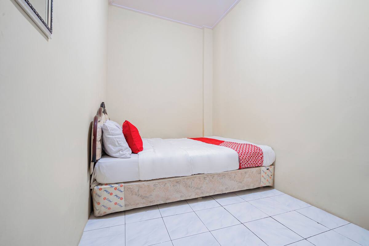 Une petite chambre avec un lit simple, un matelas blanc, une literie rouge et blanche, et un oreiller à motifs.