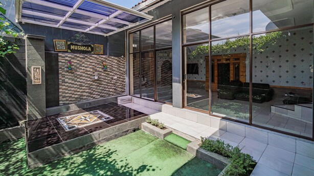 Een modern gebouw met een glazen gevel, een bakstenen muur met het bord 'MUSHOLA' en een klein terras met een patroonvloer.